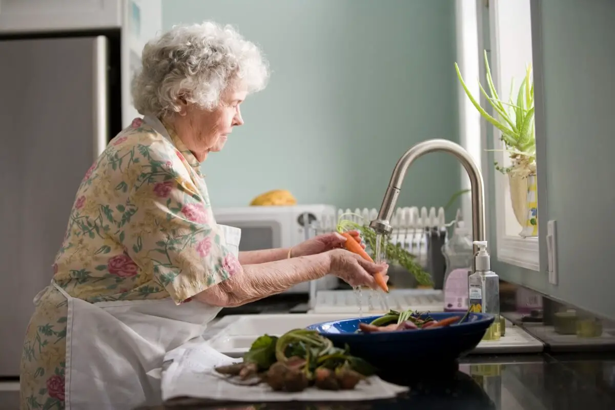 An elderly woman standing at the sink washing produce