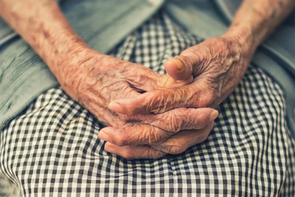 A close up image of a woman, only showing her wrinkled forearms and hands folded together and resting on her lap, which is covered in a brown and white checkered fabric.