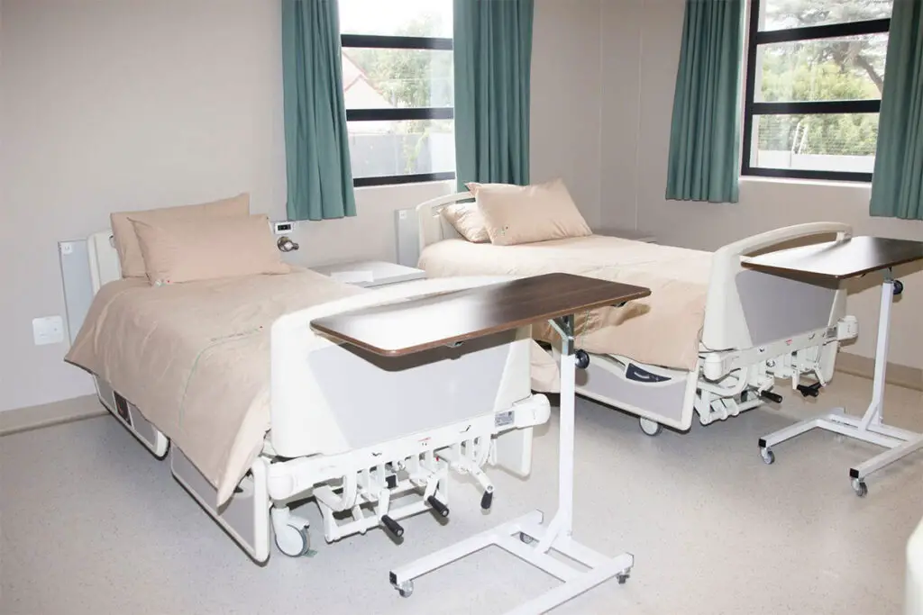 a hospital room with two beds and a desk and two overbed tables with wheels