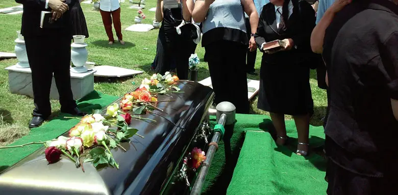 A group of people are gathered around a black coffin, standing solemnly in the grass. The majority of them are wearing dark clothing, with some dressed in suits and others wearing more casual attire.