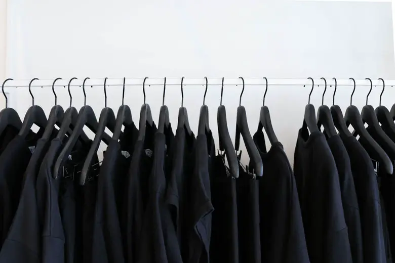 A dozen or more black clothing garments on black hangers hanging on a single white rod with a stark white backdrop.