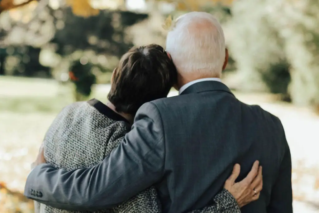 An example of appropriate choices for what to wear to a wake. A photo taken behind an aging male and female in a side embrace, standing outside in nature with their heads pressed together, the male in a gray suit and the female in a brownish gray coat. 