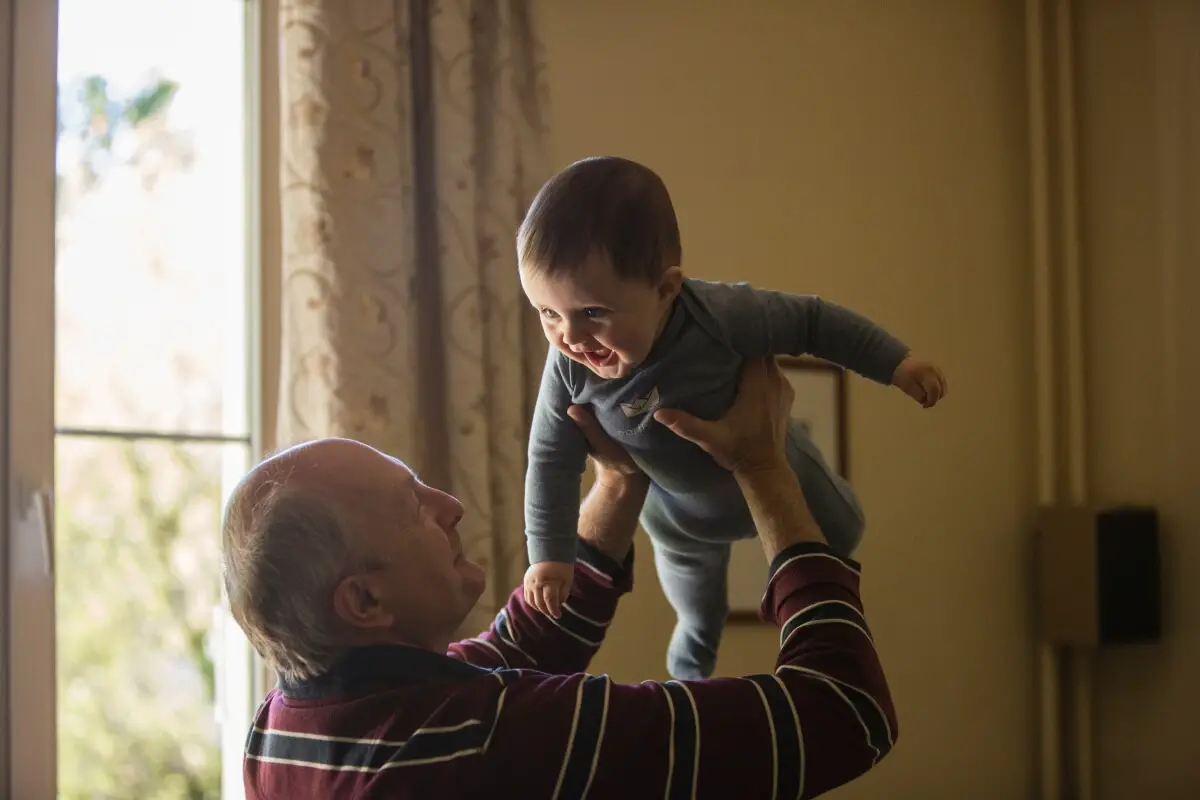 an older man playfully lifting up a young baby while wearing affordable fall protection via a medical alert device such as a fall detection button hidden beneath his clothes