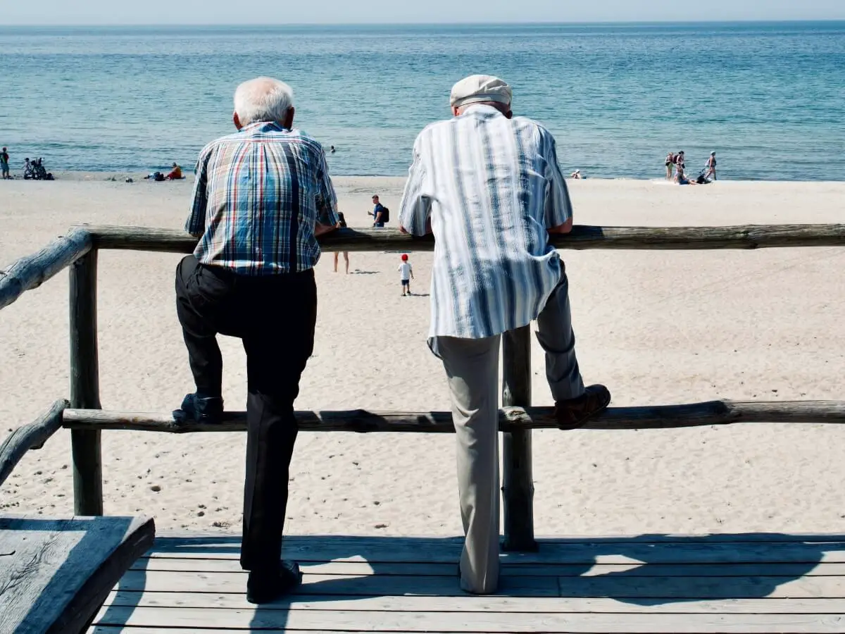 two older males standing against a railing, looking out at the ocean while comfortably wearing devices to automatically detect falls, sould they occur while either in home or on the go