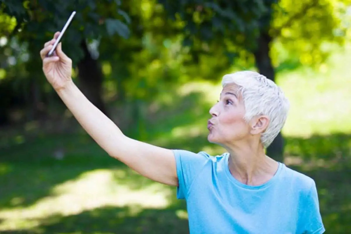 a wide eyed senior woman taking a good selfie in the park with natural light and some tree shadows in the background